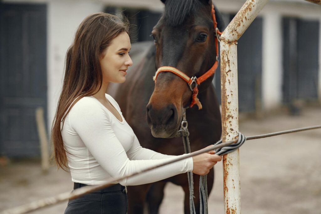 Young woman tying a horse to a steel bar outdoors, side view, smiling.